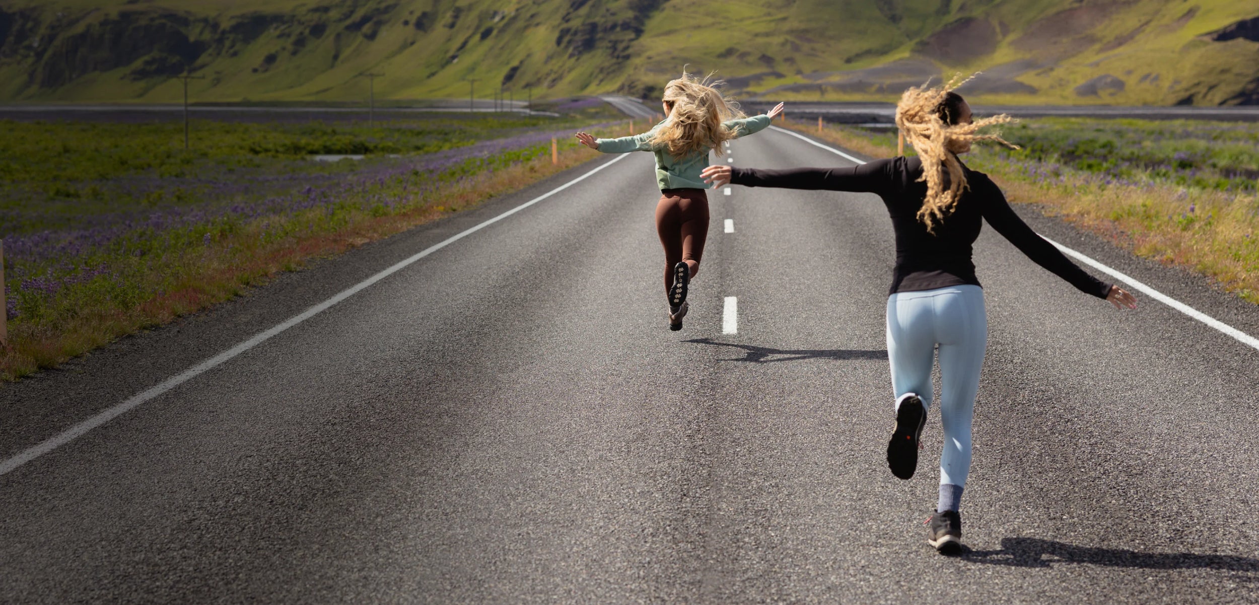 Two women running hand in hand down a paved road between green hills, viewed from behind. One wears a light green top and dark sweetflexx leggings, and the other wears a black long-sleeve top and light blue leggings.